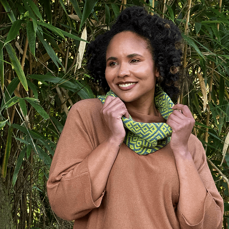 Smiling woman in a tan shirt wearing the Encaustic cowl in slate blue and citronella, in front of a stand of bamboo